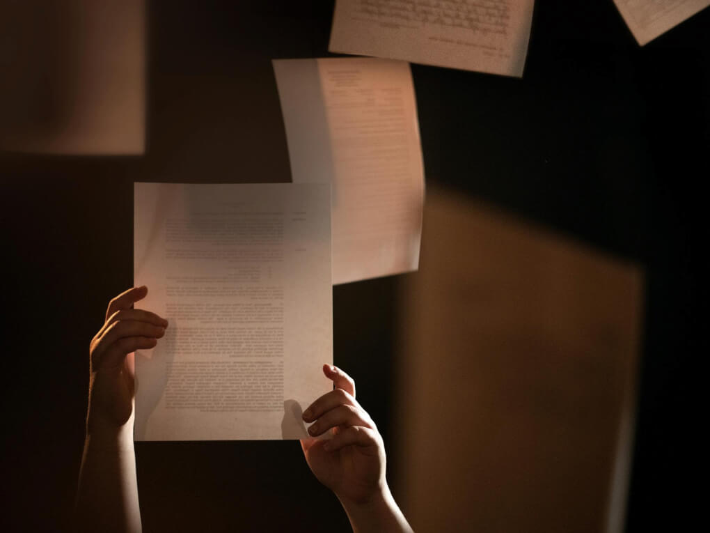 Two hands grasp a piece of paper that appears to be floating in the air, other papers float around it. There's blurred writing and type on some of the papers. The image is dramatically lit. The papers represent evidence in a cold case class.